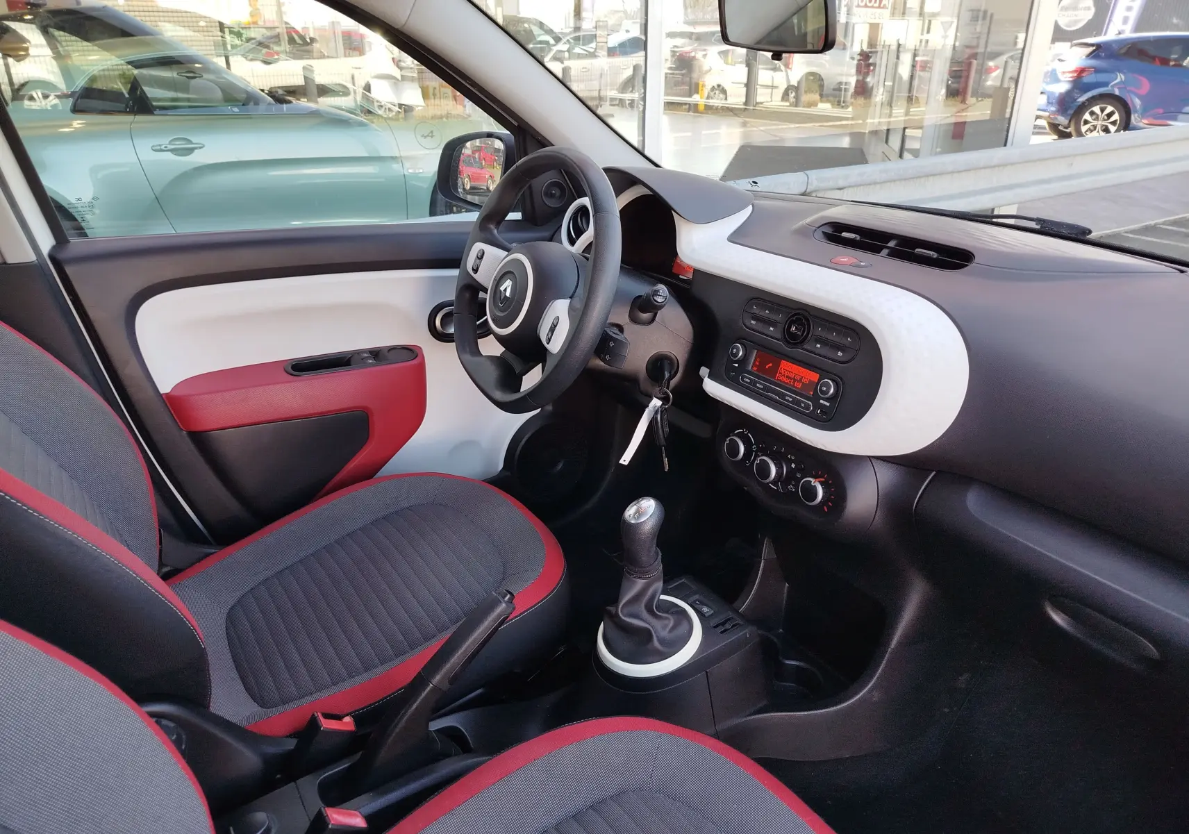 Vue intérieure côté conducteur de la Renault Twingo 2016, tableau de bord blanc texturé et sièges gris avec surpiqûres rouges.