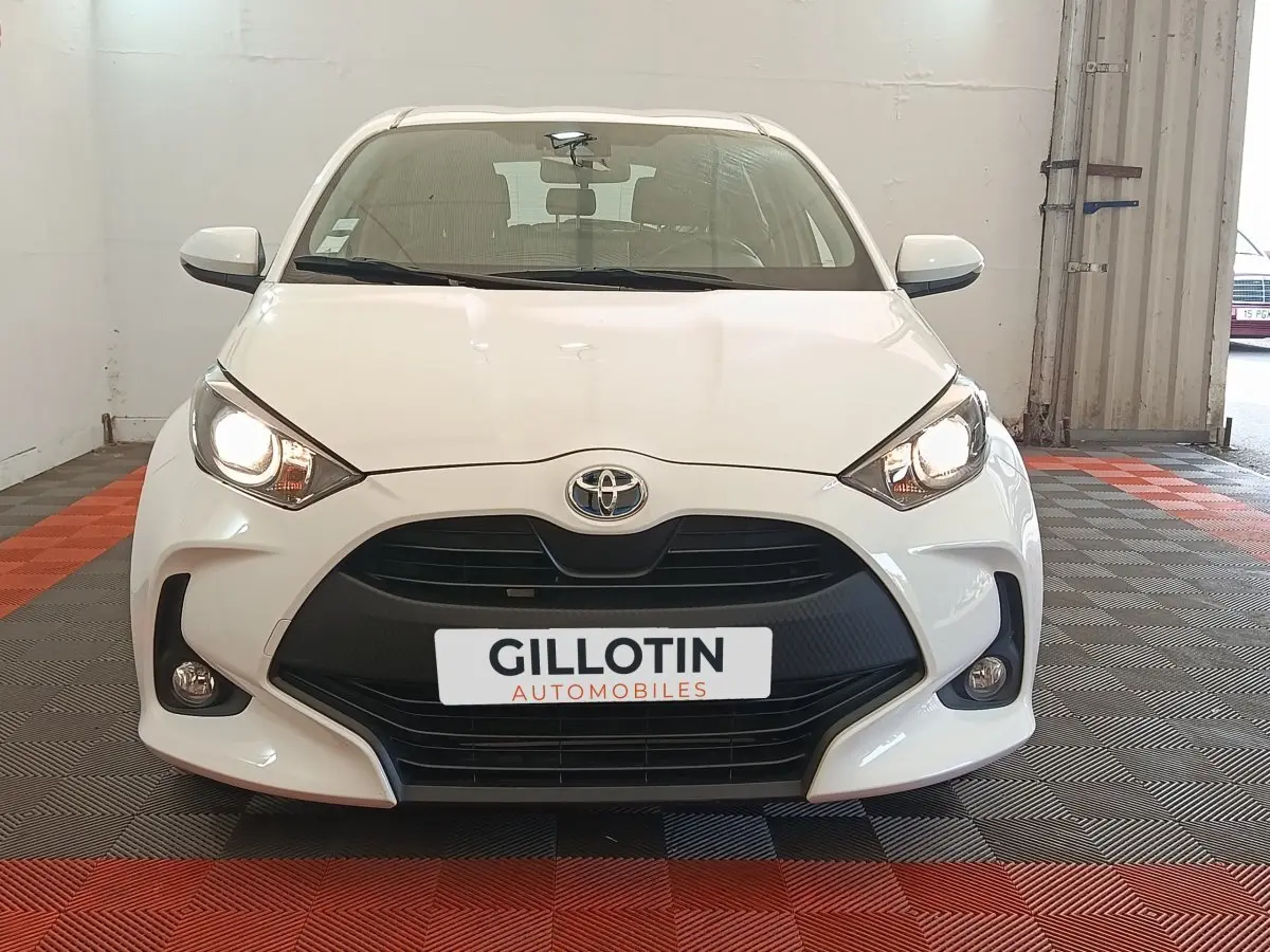 Vue frontale d'une Toyota Yaris hybride blanche avec phares allumés et calandre noire dans un showroom.