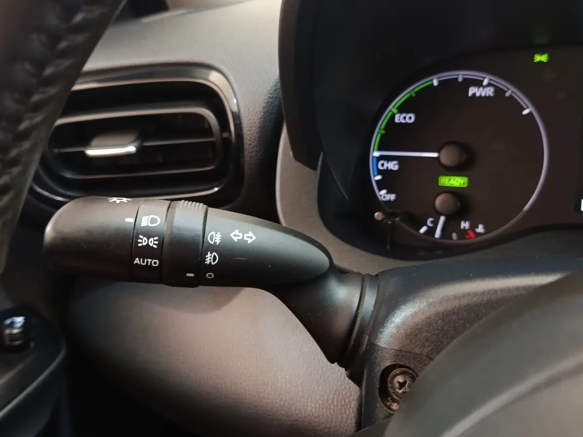 Gros plan sur la manette de commande des phares et clignotants côté gauche du tableau de bord noir de la Toyota Yaris hybride blanche.