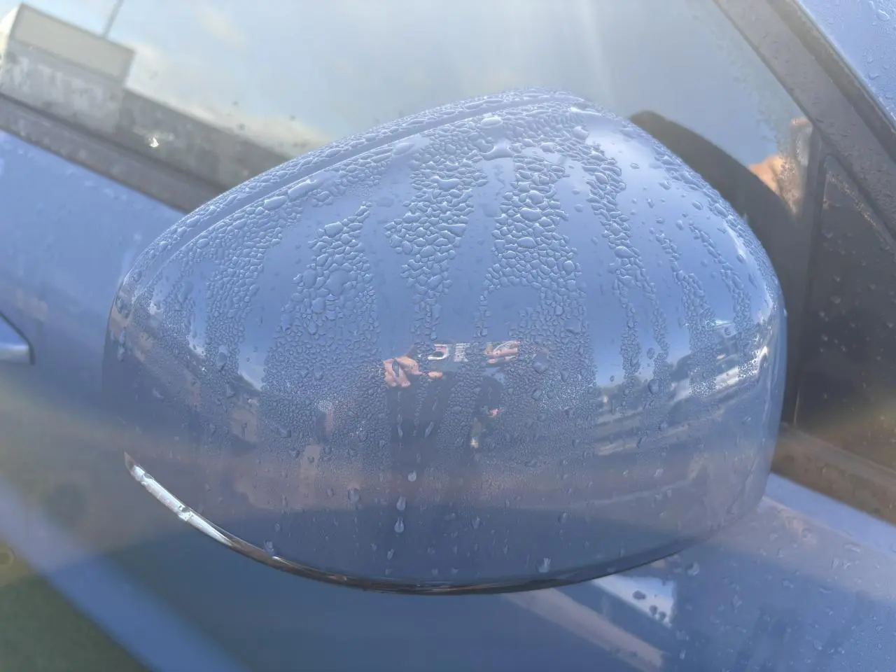 Rétroviseur côté gauche bleu Fjord Blue métallisé du Volvo XC40 2024 avec gouttes d'eau visibles.