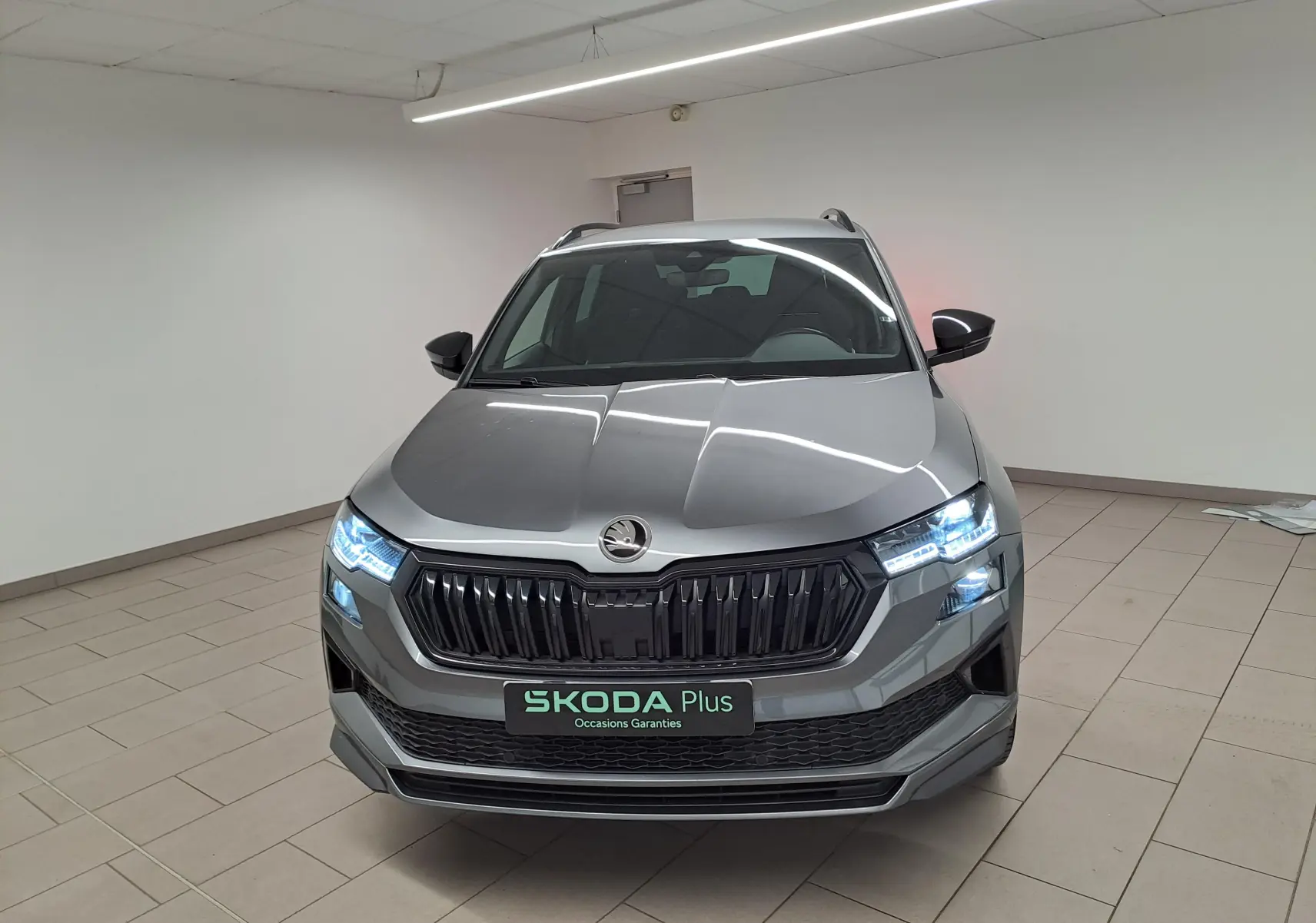 Vue avant d'un Skoda Karoq 2.0 TDI gris graphite avec phares allumés dans un showroom intérieur.