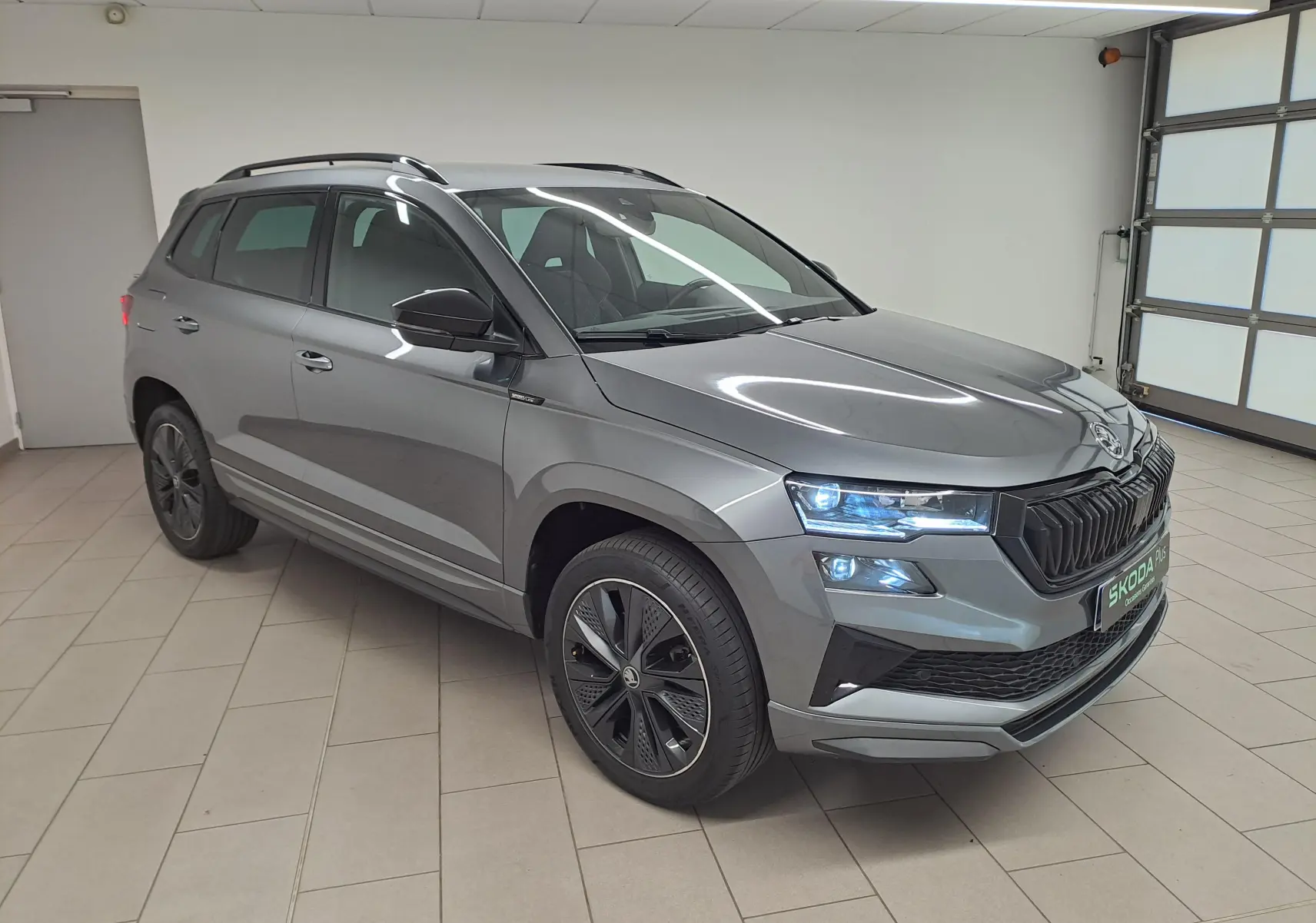Vue 3/4 avant droite du Skoda Karoq gris graphite 2025 avec phares allumés et jantes noires sportline.