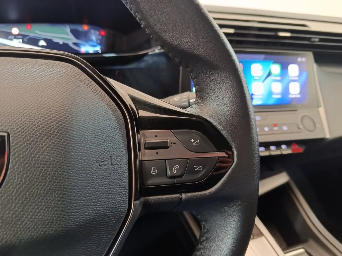 Gros plan sur le volant cuir noir côté droit de la Peugeot 308 2023 avec commandes vocales et écran tactile en arrière-plan.