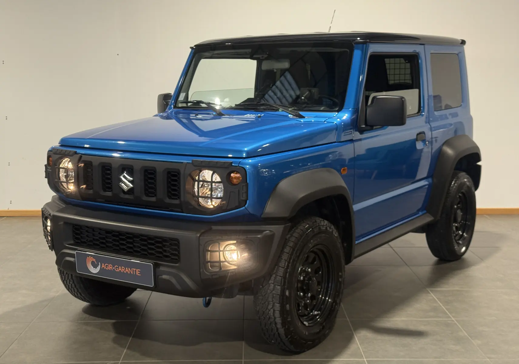 Suzuki Jimny 1.5 VVT bleu en 3/4 avant droit, avec phares et antibrouillards allumés, finition Privilège Allgrip.