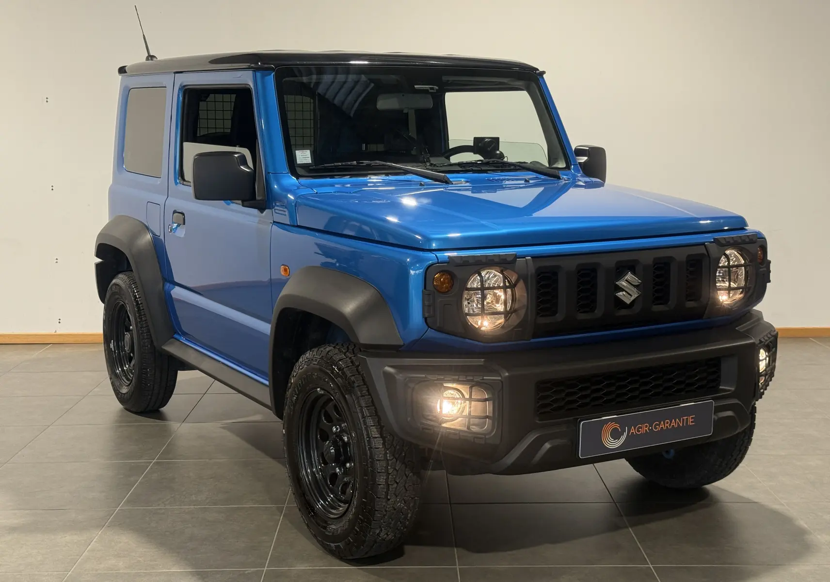 Suzuki Jimny 2021 bleu en 3/4 avant droit, avec phares et antibrouillards allumés, toit noir et protections noires.