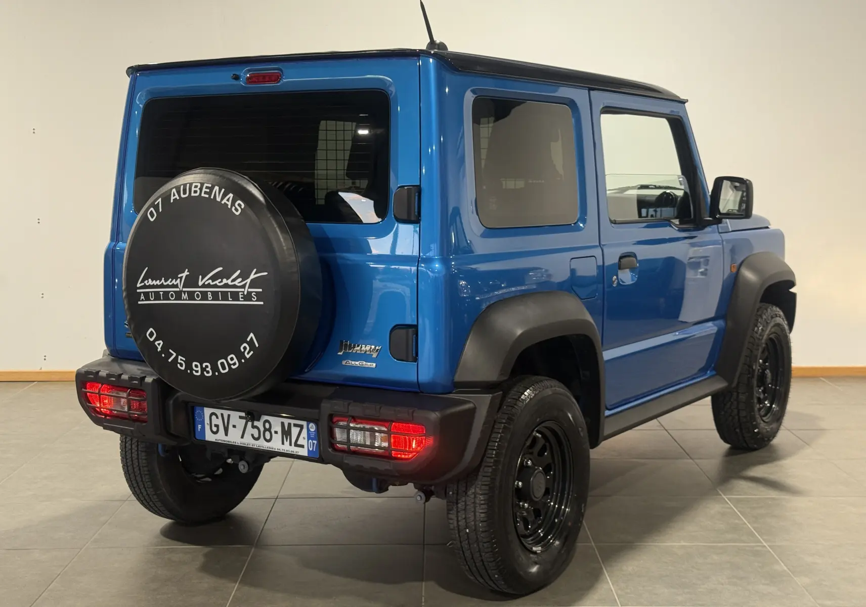 Vue 3/4 arrière droite d'un Suzuki Jimny bleu 2021 avec roue de secours noire et protections d'ailes noires.