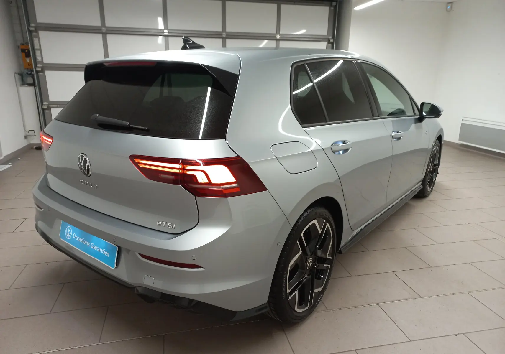 Vue 3/4 arrière droit d'une Volkswagen Golf 1.5 eTSI argent dolomite avec jantes noires et feux LED allumés.