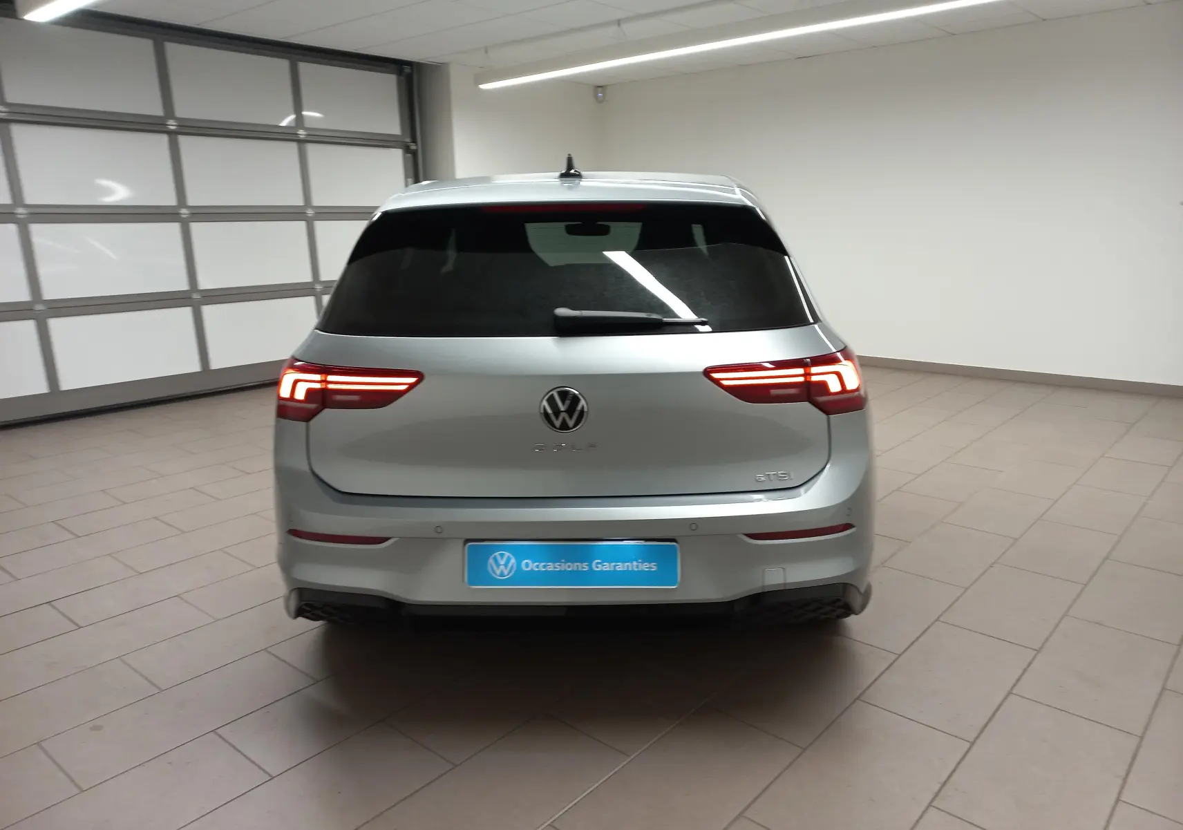 Vue arrière d'une Volkswagen Golf 1.5 eTSI 150 DSG7 R-Line Edition en argent dolomite avec feux LED allumés dans un garage.