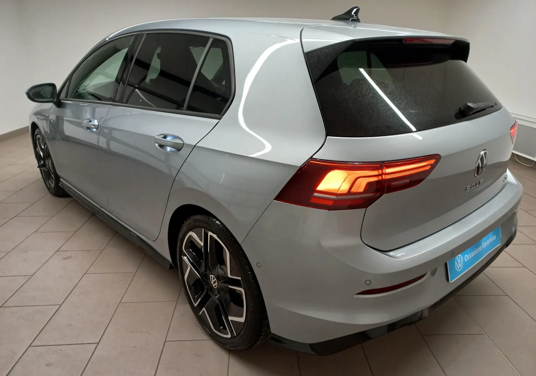 Vue 3/4 arrière gauche d'une Volkswagen Golf 1.5 eTSI R-Line argent dolomite avec feux allumés et jantes spécifiques.