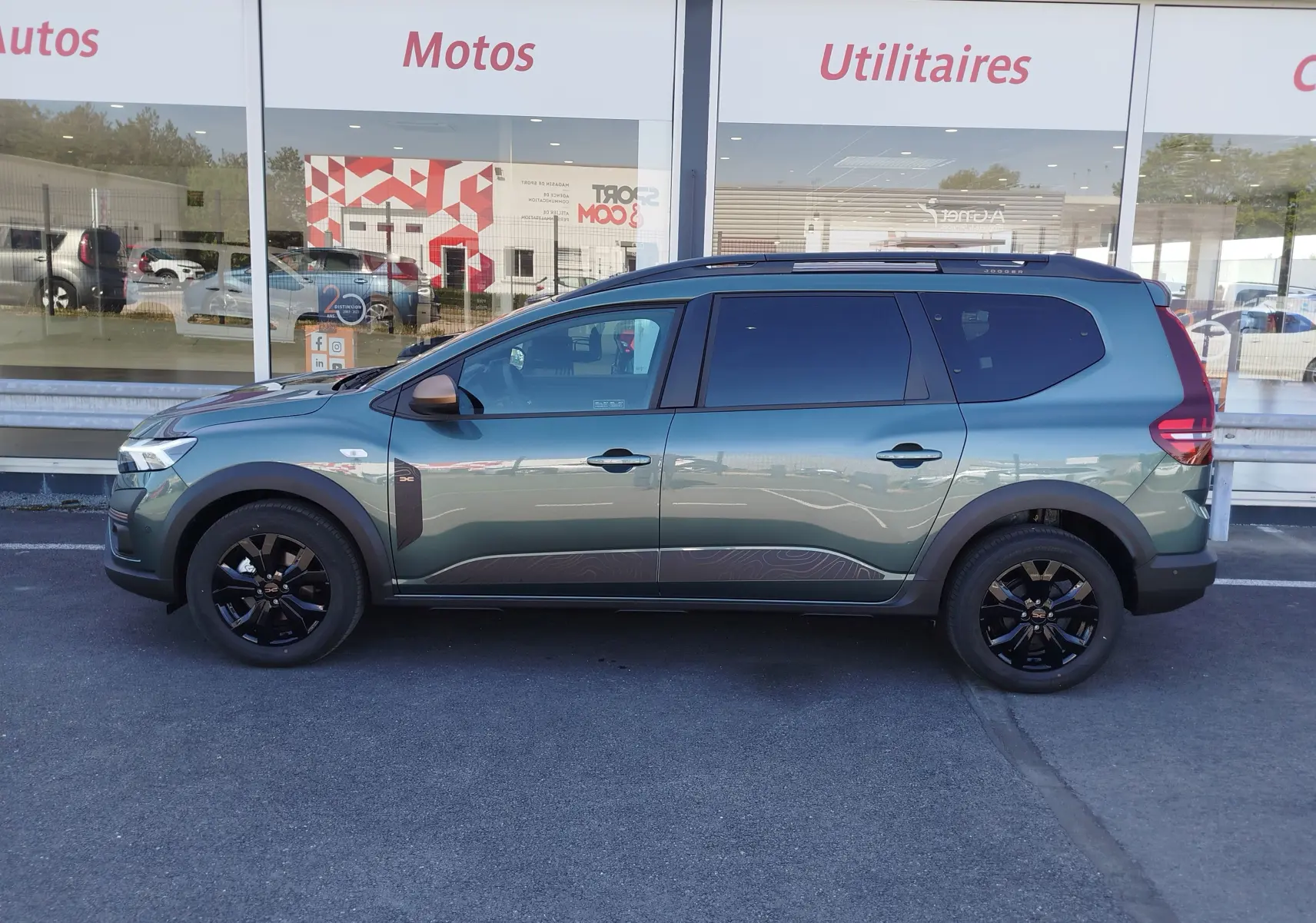 Profil droit d'un Dacia Jogger 2026 vert cèdre métal avec jantes noires et barres de toit, stationné devant une vitrine.