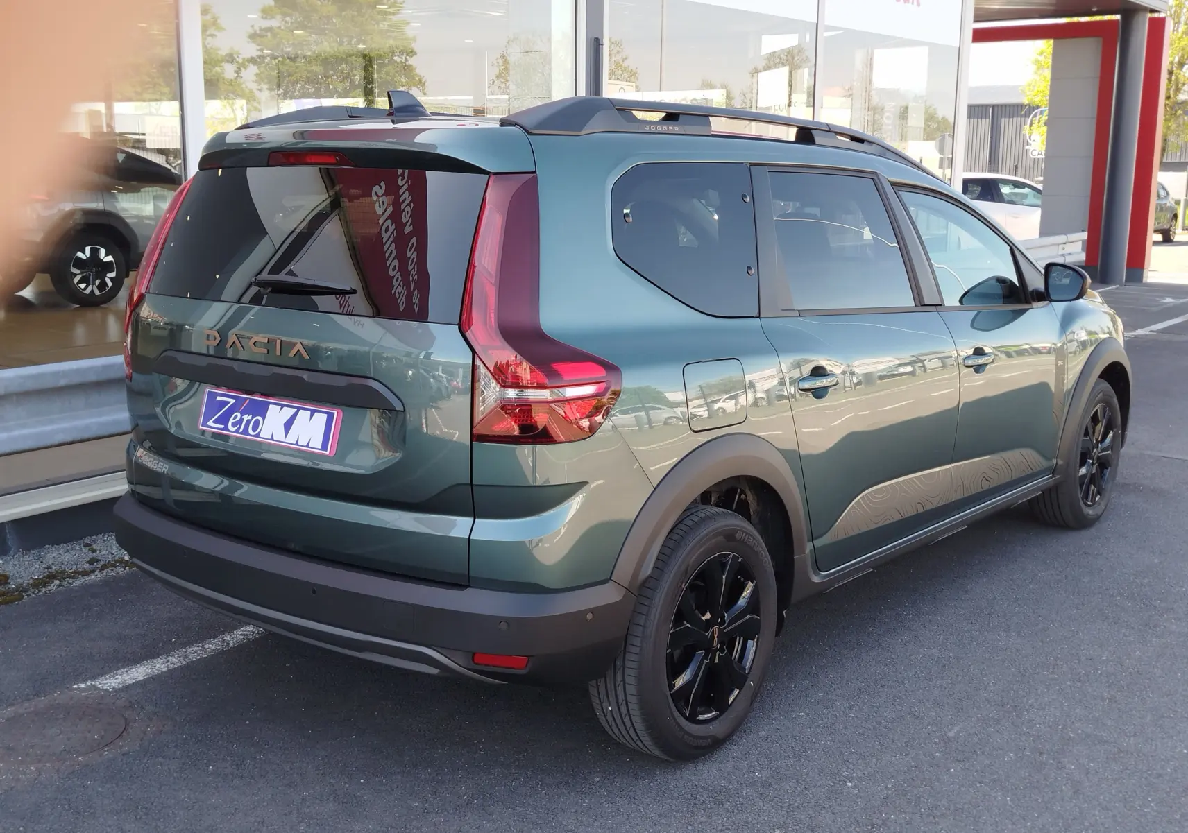 Vue 3/4 arrière droite d'un Dacia Jogger vert cèdre métallisé avec jantes noires et barres de toit, stationné devant un showroom.