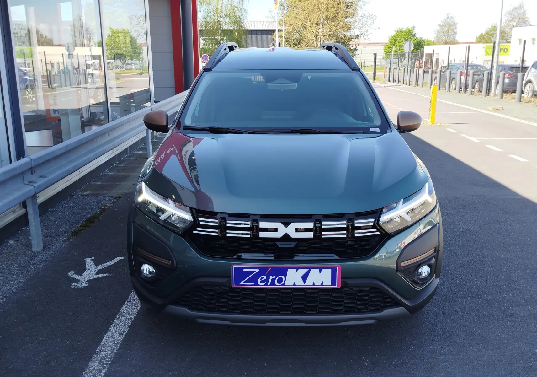 Vue avant d'un Dacia Jogger 2026 vert cèdre métal avec calandre noire et phares LED allumés, stationné devant un bâtiment.