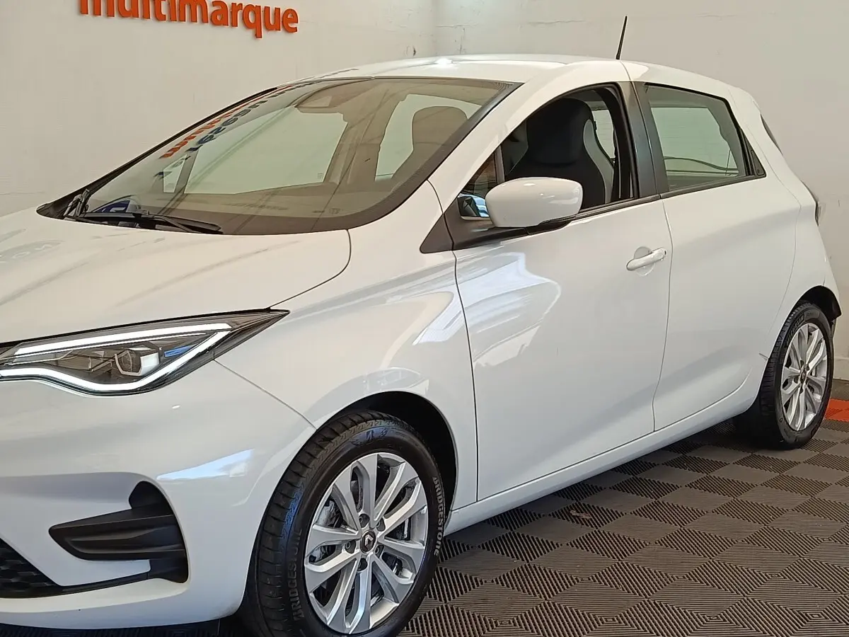Renault ZOE blanc vue 3/4 avant droit, phares LED et jantes alliage visibles en intérieur showroom.
