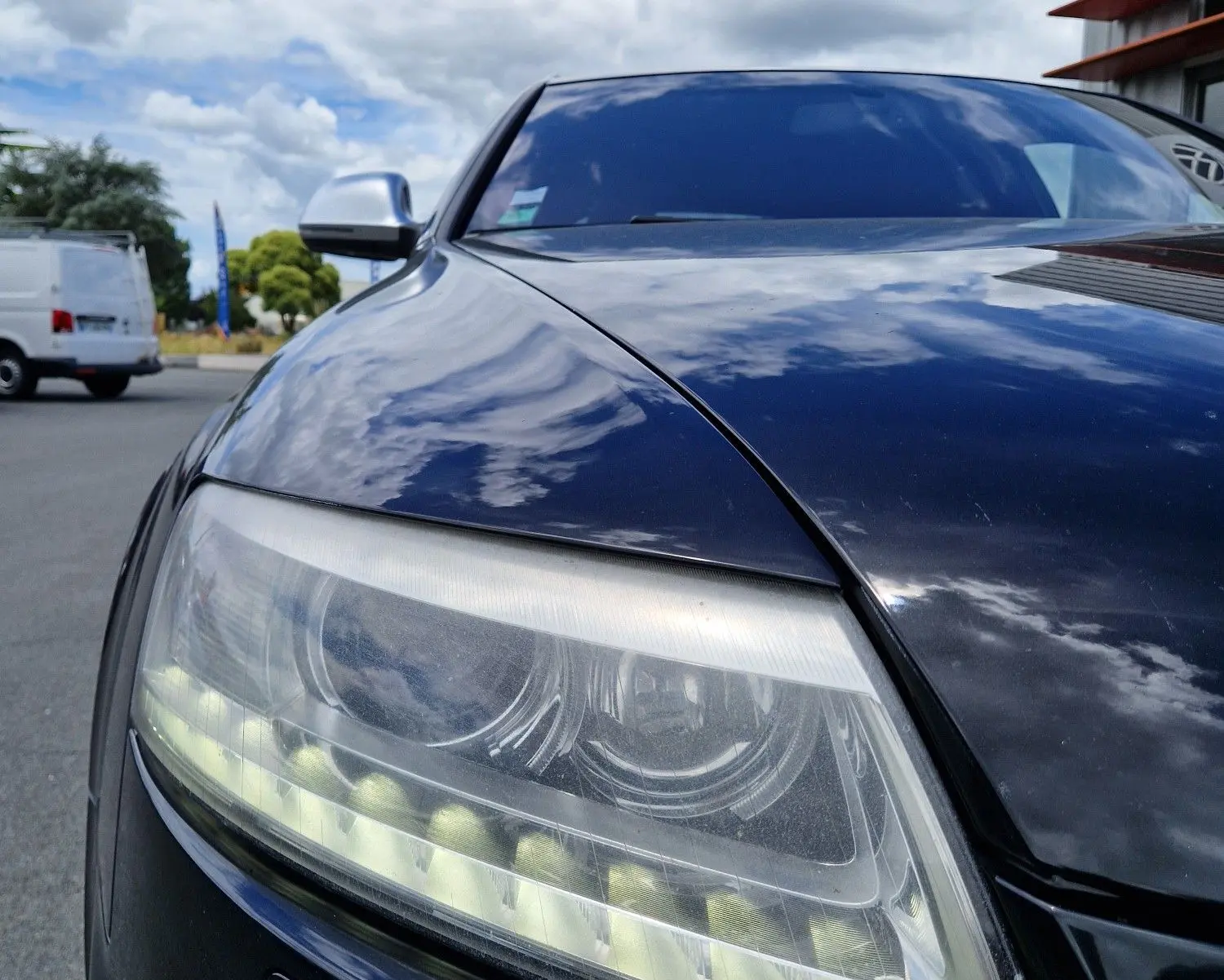 Gros plan sur le phare avant gauche noir brillant d'une Audi RS6 Avant 2009, reflétant le ciel nuageux.