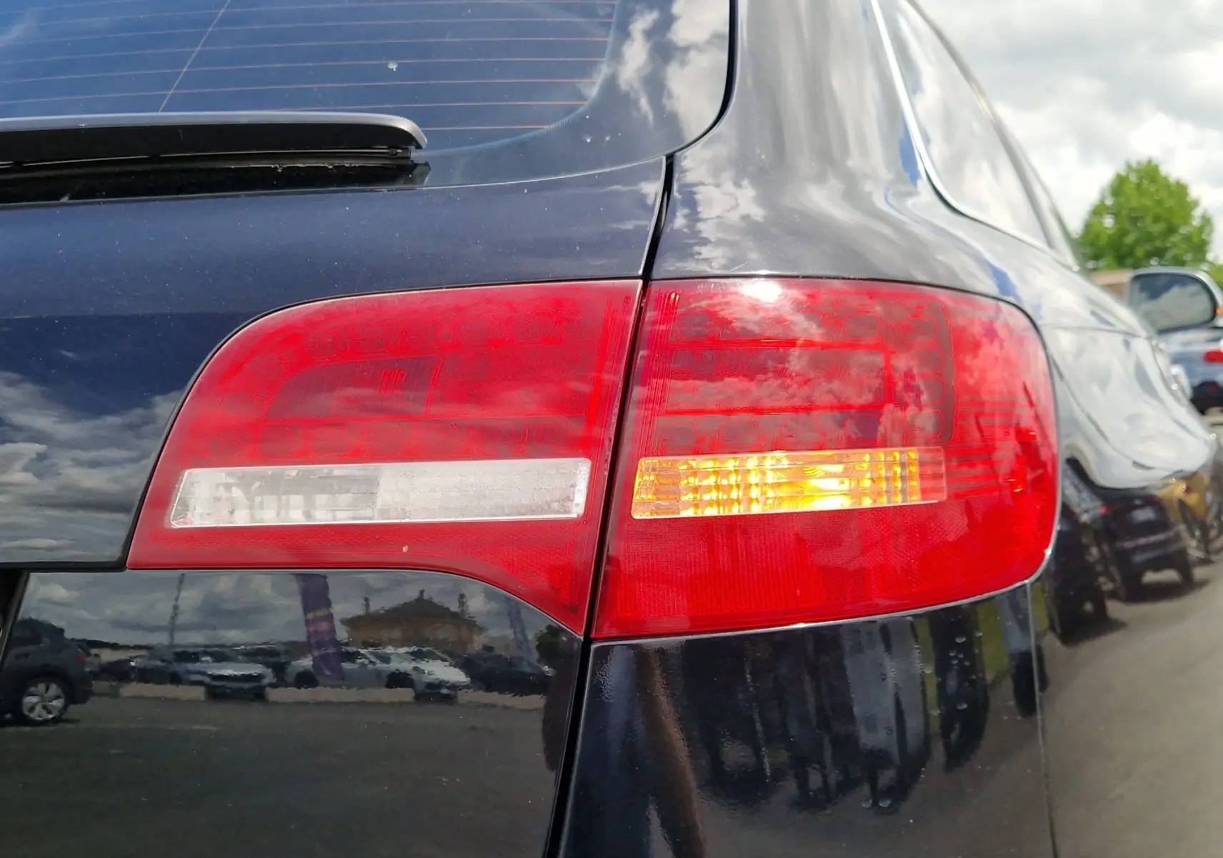 Gros plan sur le feu arrière droit rouge et orange d'une Audi RS6 Avant noire, reflétant le ciel et le parking environnant.