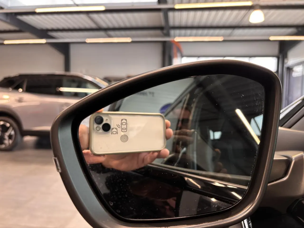 Rétroviseur extérieur droit noir brillant du Peugeot 2008 blanc, reflétant partiellement l'intérieur et un smartphone.