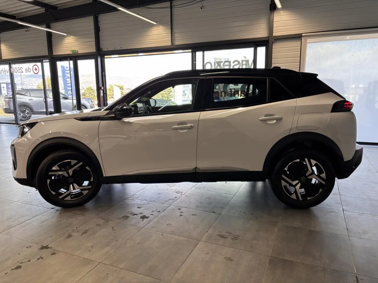 Vue de profil côté gauche d’un Peugeot 2008 blanc avec toit noir et jantes alliage bi-ton dans un showroom.