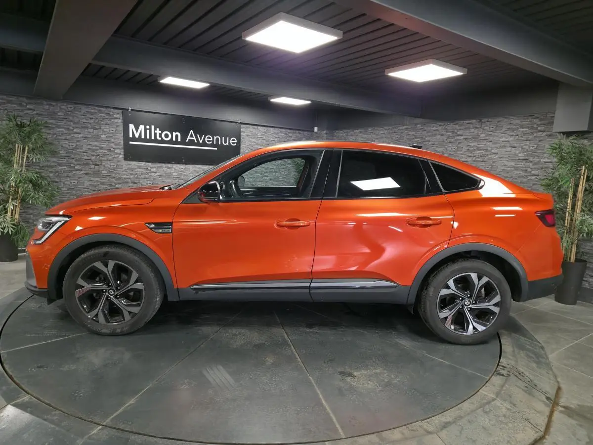 Profil droit du Renault Arkana 2024 orange Valencia avec jantes 18 pouces et toit ouvrant électrique visible en intérieur showroom.