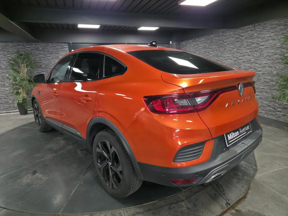 Vue 3/4 arrière droite d'un Renault Arkana orange Valencia avec jantes noires et toit ouvrant en intérieur showroom.