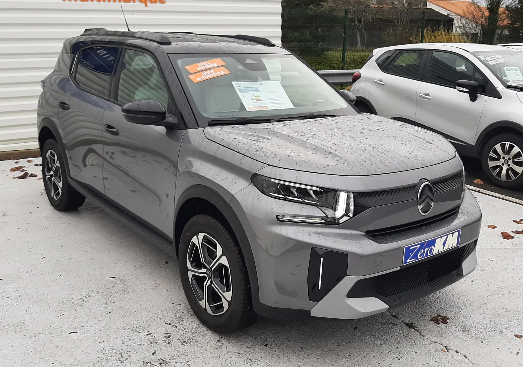 Citroën C3 Aircross hybride gris Mercury avec toit noir, vue 3/4 avant droit sous la pluie sur parking multimarque.
