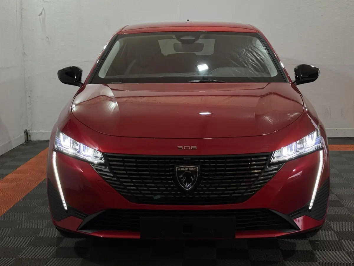 Vue frontale d'une Peugeot 308 rouge 2025 avec phares LED allumés et calandre noire distinctive.
