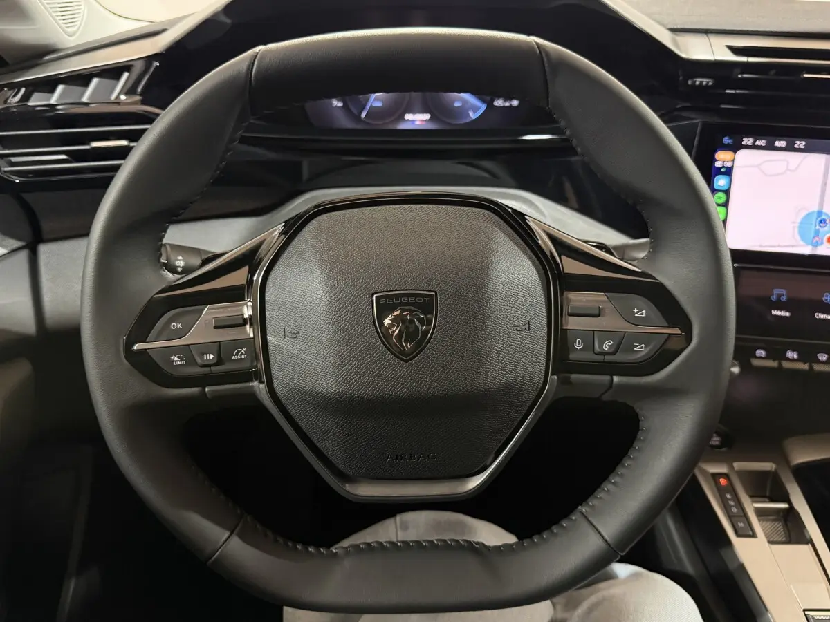 Vue rapprochée du volant cuir noir de la Peugeot 308 2025 avec commandes multifonctions et écran tactile en arrière-plan.
