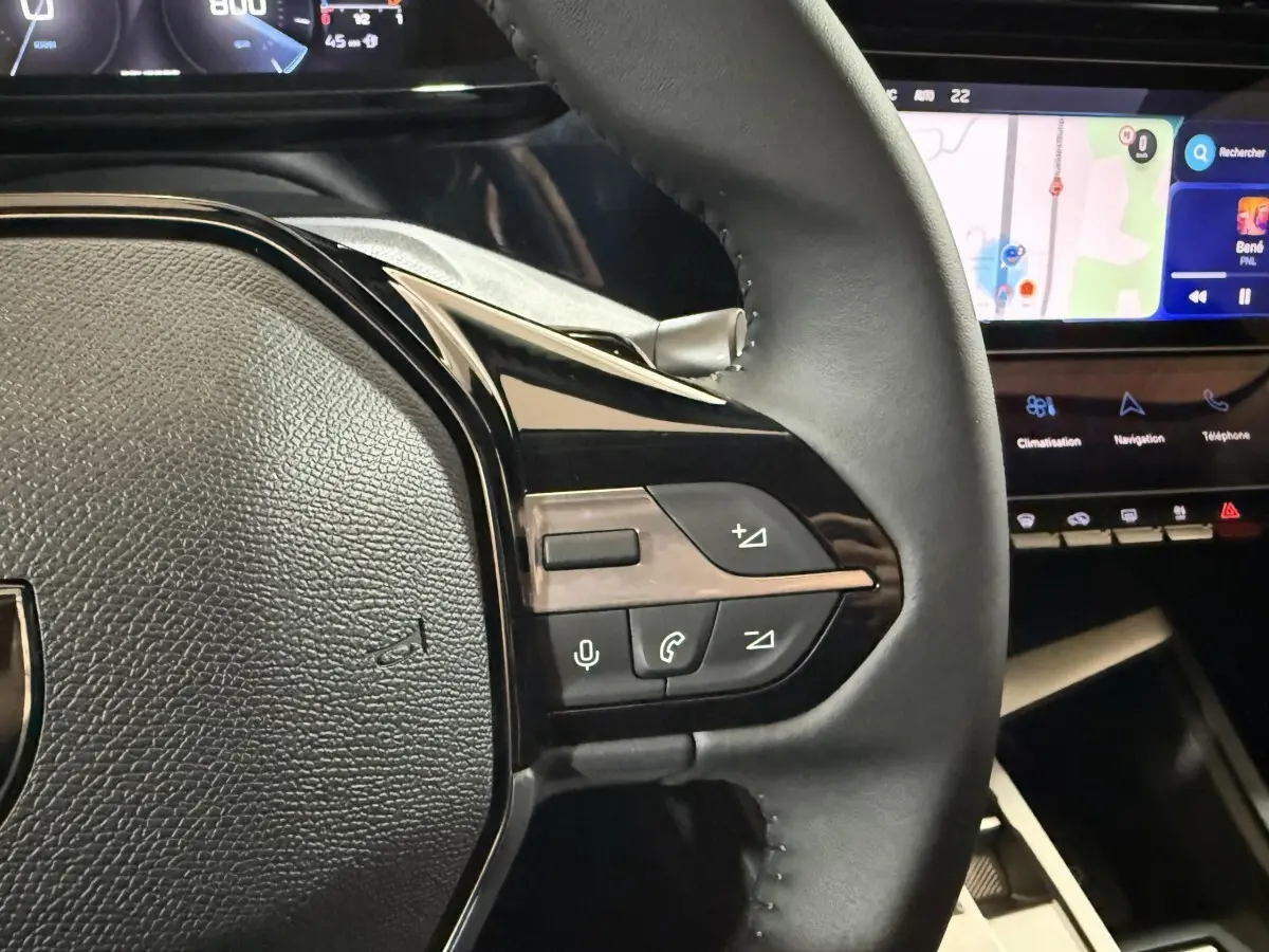 Gros plan sur le volant cuir à commandes multifonctions côté droit du Peugeot 308 rouge, avec écran tactile de navigation en arrière-plan.