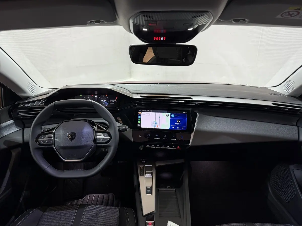 Vue intérieure avant du Peugeot 308 2025 avec tableau de bord noir, écran tactile central et volant cuir multifonctions.