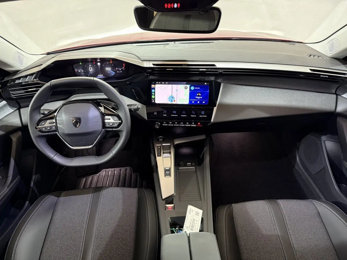 Vue intérieure avant de la Peugeot 308 2025, tableau de bord noir avec écran tactile central et volant multifonctions cuir.