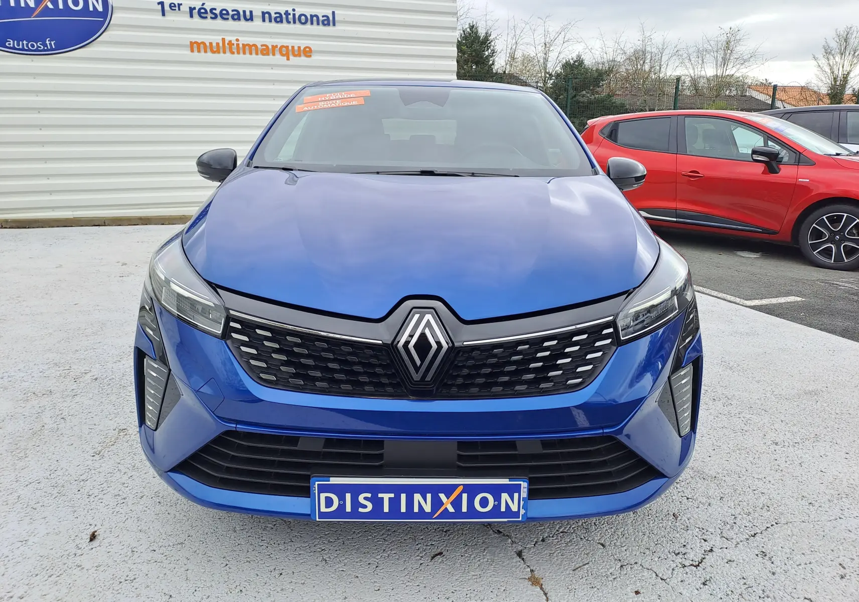 Vue frontale d'une Renault Clio V bleu Iron Metal avec calandre noire et logo Renault proéminent, stationnée en extérieur.