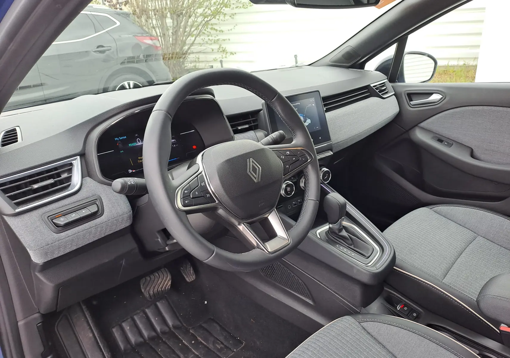 Vue intérieure côté conducteur de la Renault Clio V hybride 2025, tableau de bord et volant noirs avec sellerie grise.