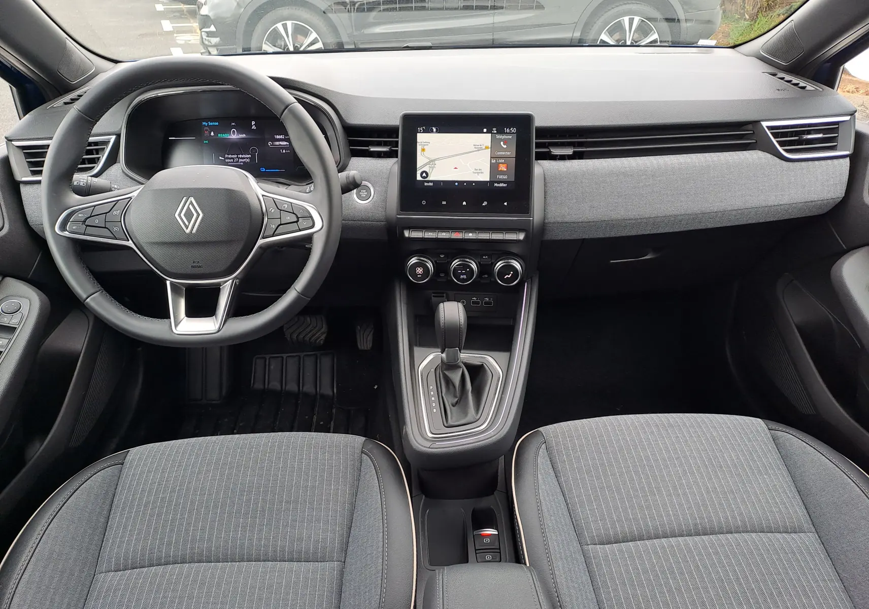 Vue intérieure avant de la Renault Clio V 1.6 E-TECH avec tableau de bord moderne, écran tactile central et sièges tissu gris.