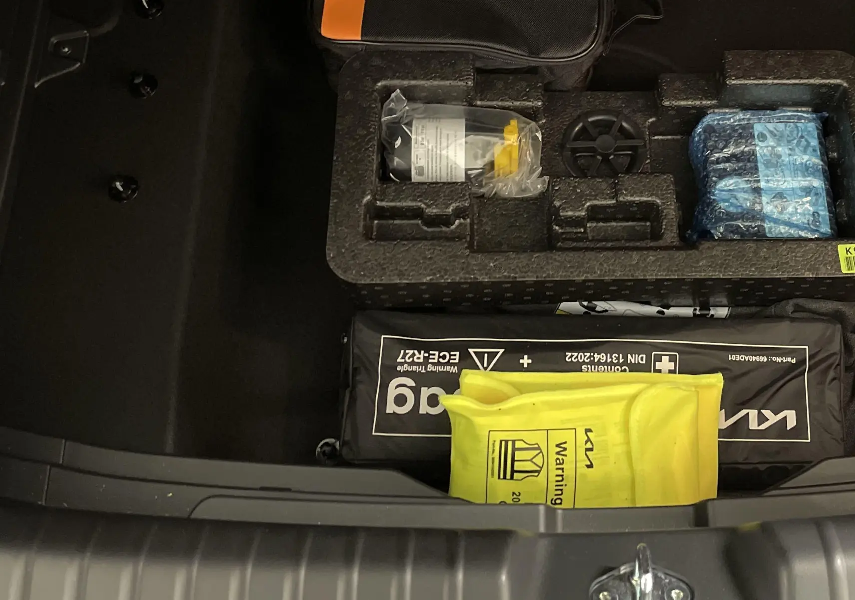 Coffre ouvert du Kia Niro blanc 2025 montrant le rangement sous plancher avec trousse de secours et kit de sécurité.