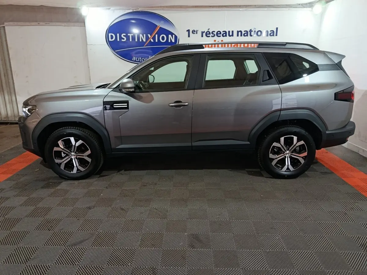 Vue de profil côté gauche d'un Dacia Bigster gris foncé avec jantes alliage et barres de toit noires.