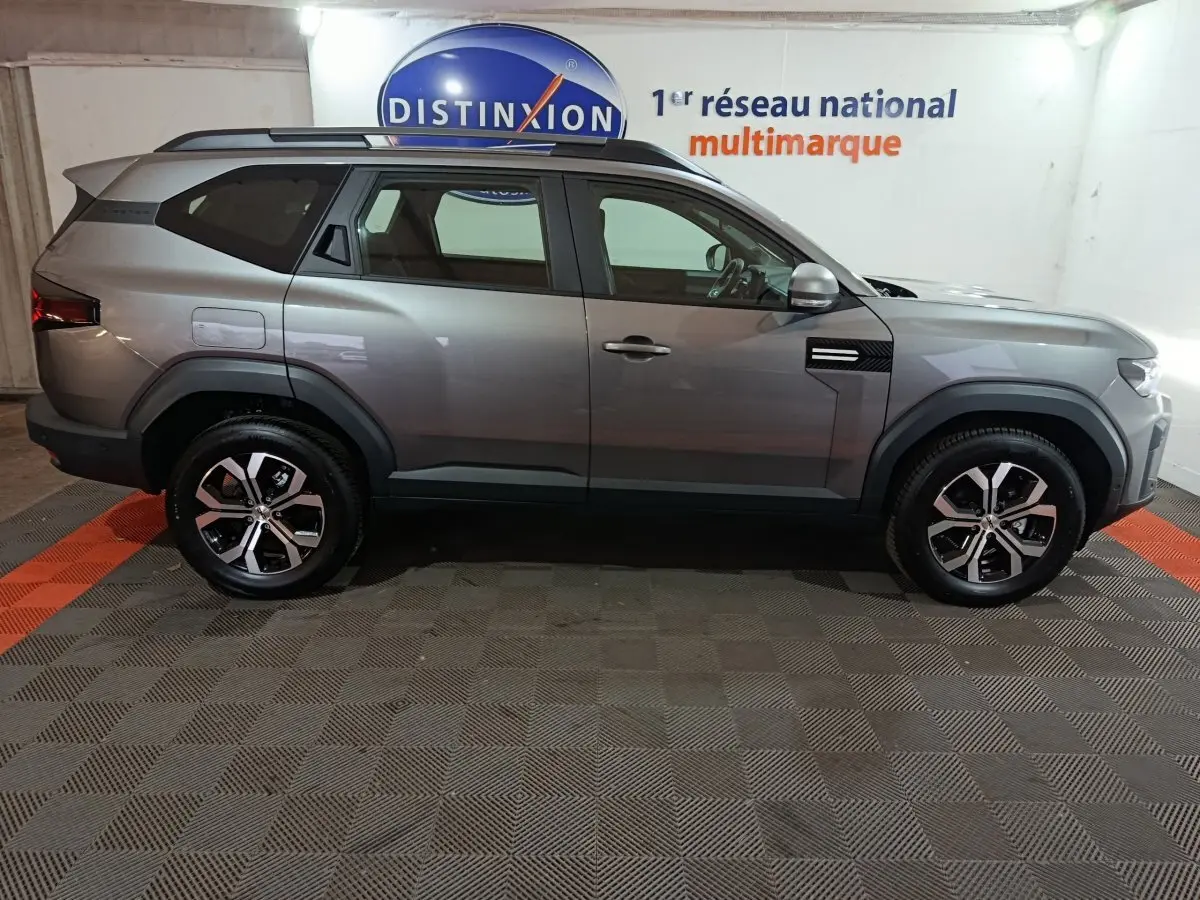 Vue de profil droit du SUV Dacia Bigster gris foncé 2025 avec jantes alliage et barres de toit noires.