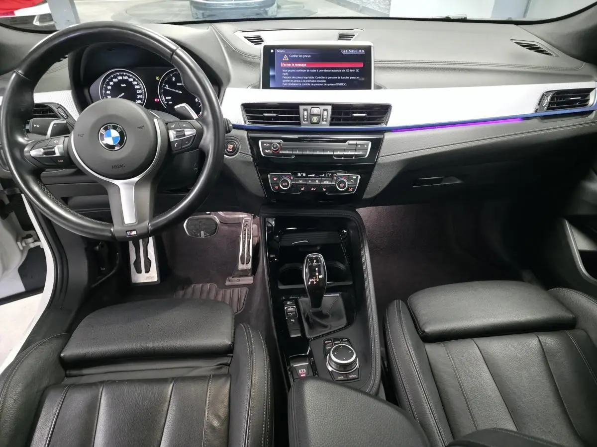 Vue intérieure avant du BMW X2 sDrive 18d M Sport 2023, avec sellerie cuir noire et éclairage d'ambiance violet sur la planche de bord.