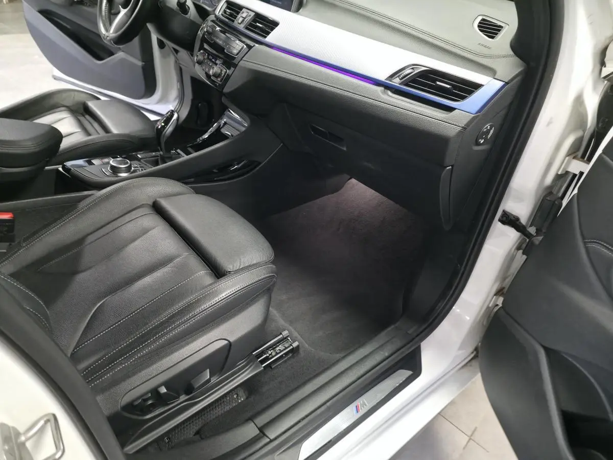 Vue intérieure côté passager avant d'une BMW X2 blanche, mettant en valeur la sellerie cuir noire et l'éclairage d'ambiance bleu.