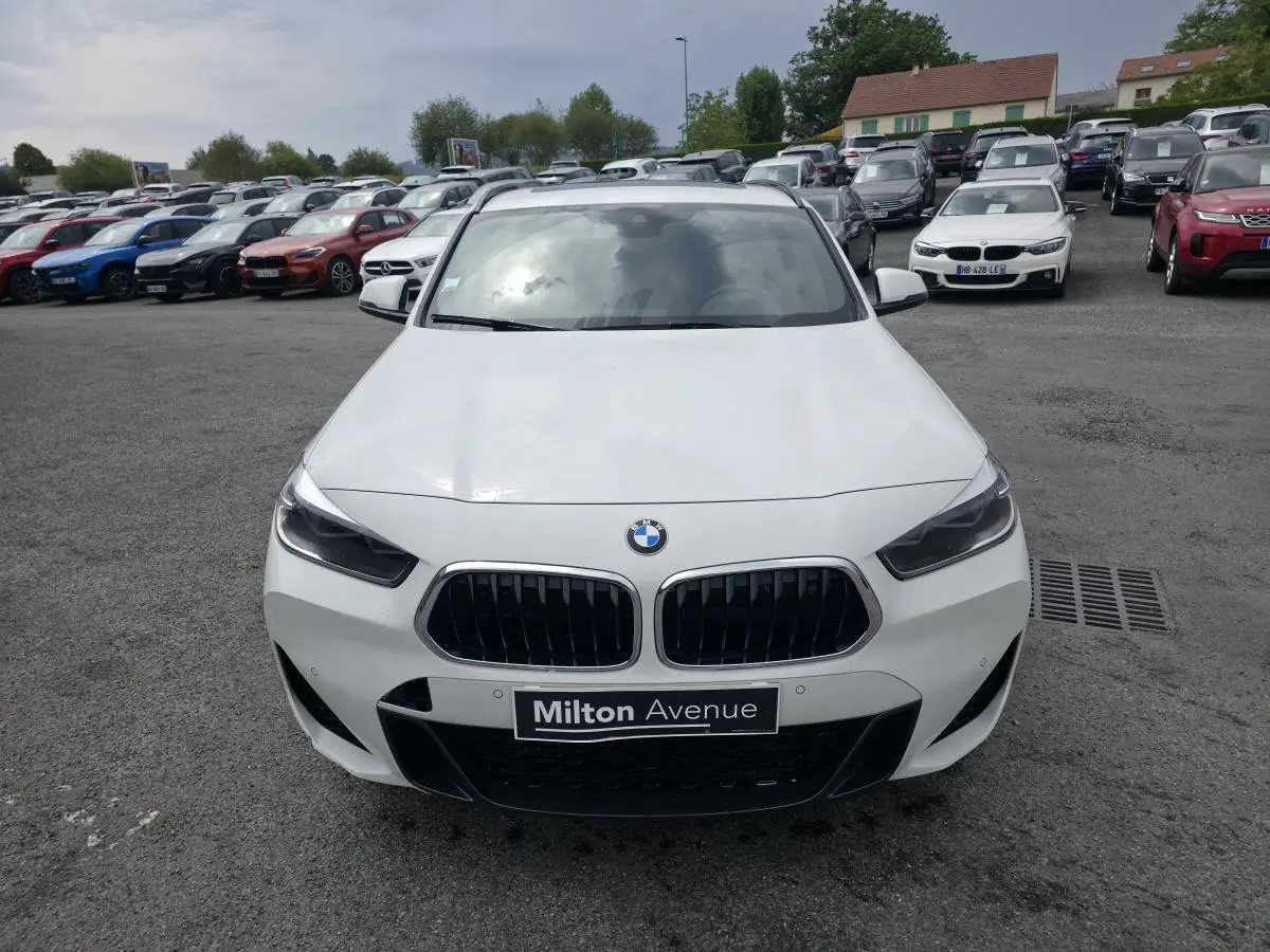 Vue avant d'une BMW X2 sDrive 18d blanche avec calandre noire et plaque Milton Avenue visible en concession.