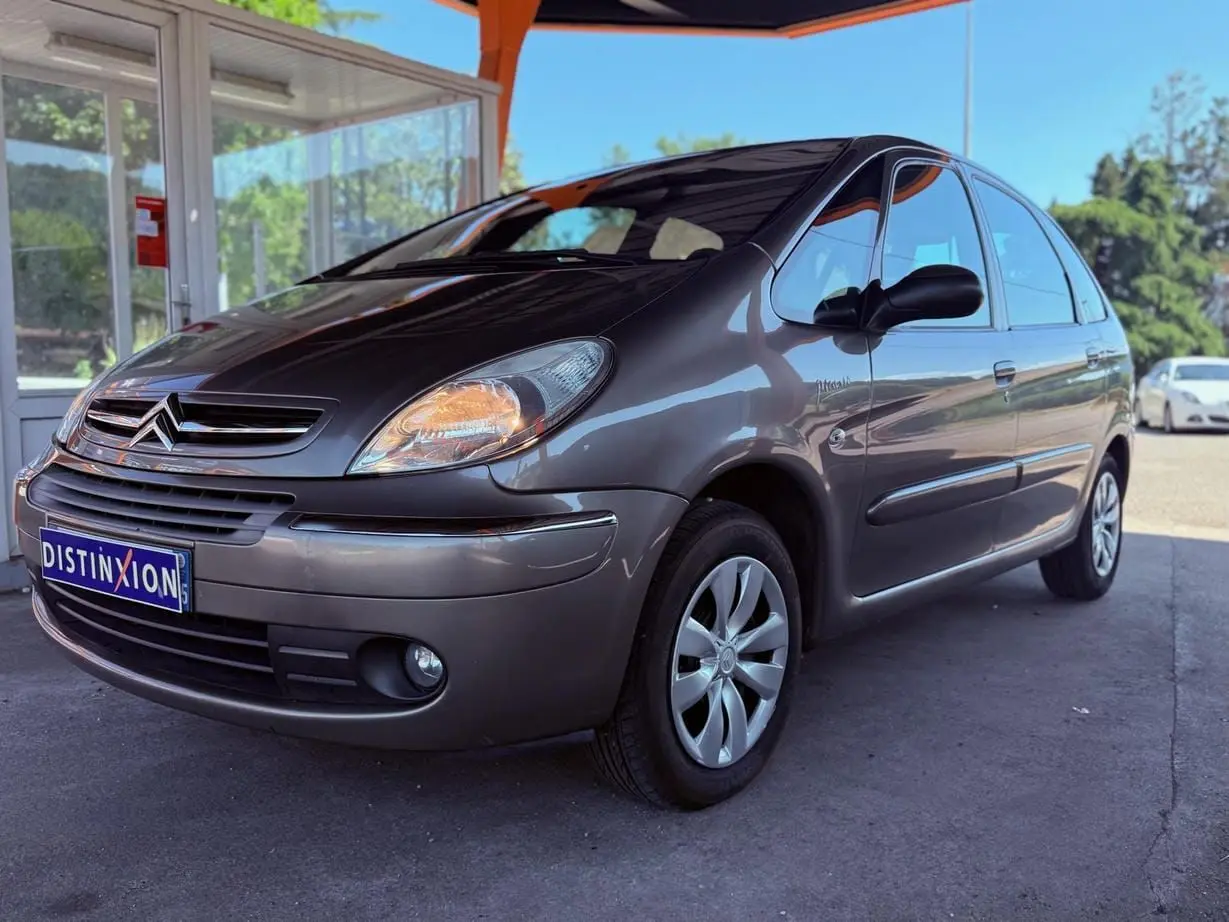 Vue 3/4 avant droit d'une Citroën Xsara Picasso beige métallisé garée sous un abri avec phares allumés.