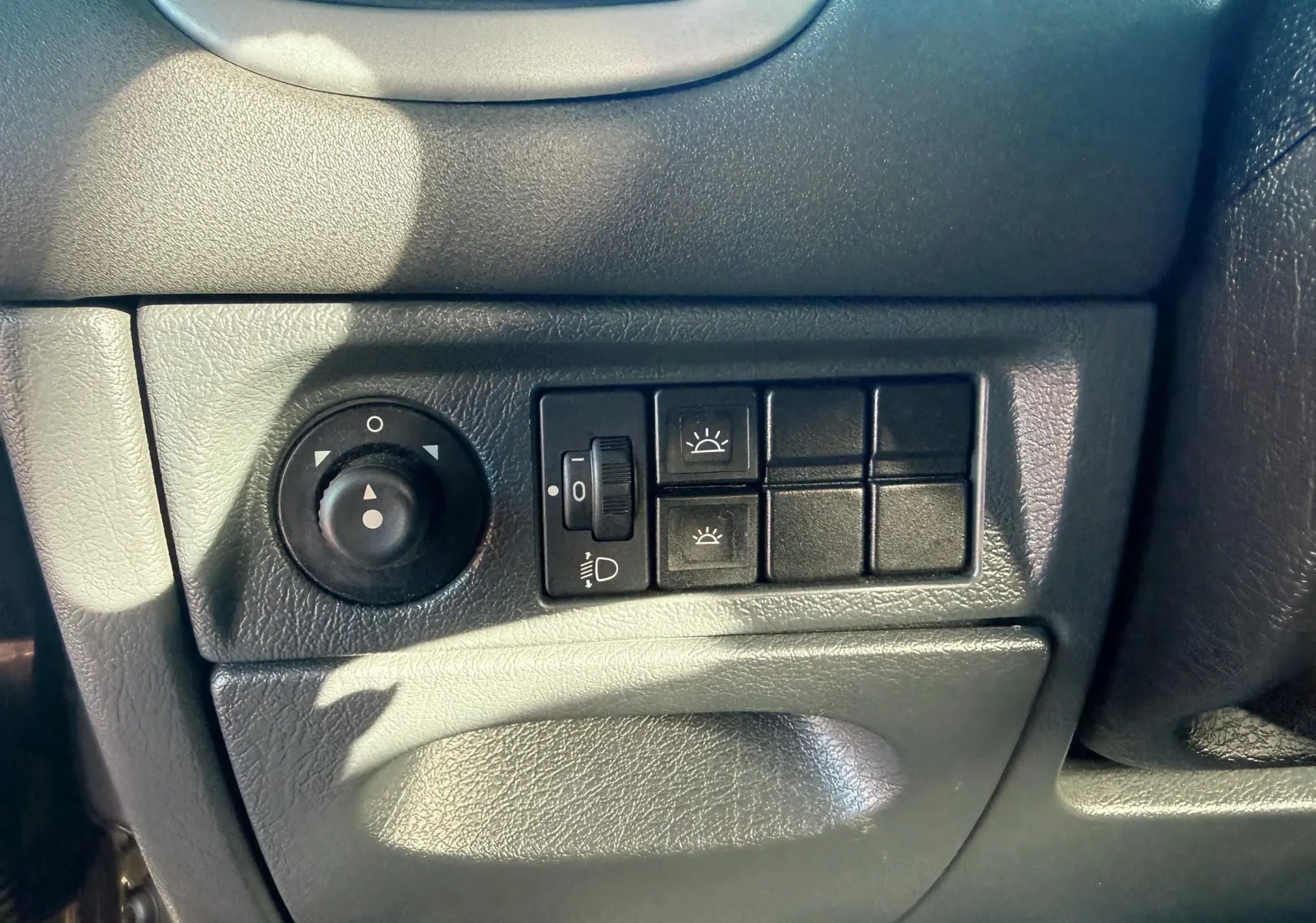 Gros plan sur les commandes d’éclairage et réglage des rétroviseurs du tableau de bord noir d’une Citroën Xsara Picasso beige.