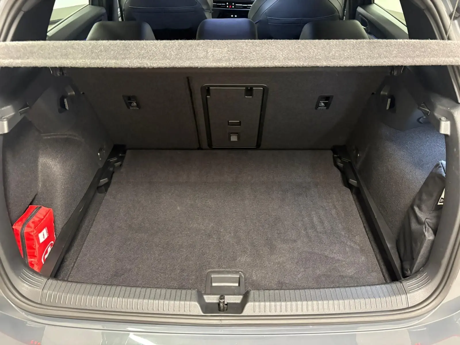 Coffre ouvert vu de l'arrière d'une Volkswagen Golf 1.5 eTSI gris dauphin, avec tapis noir et trousse de secours rouge à gauche.