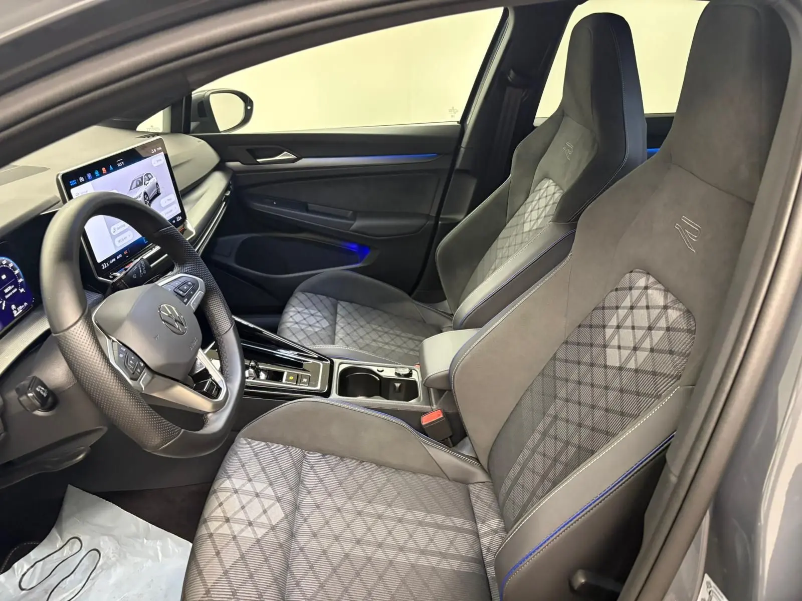 Vue intérieure côté conducteur de la Volkswagen Golf 1.5 eTSI R-Line 2025 avec sièges gris à surpiqûres bleues et écran tactile.