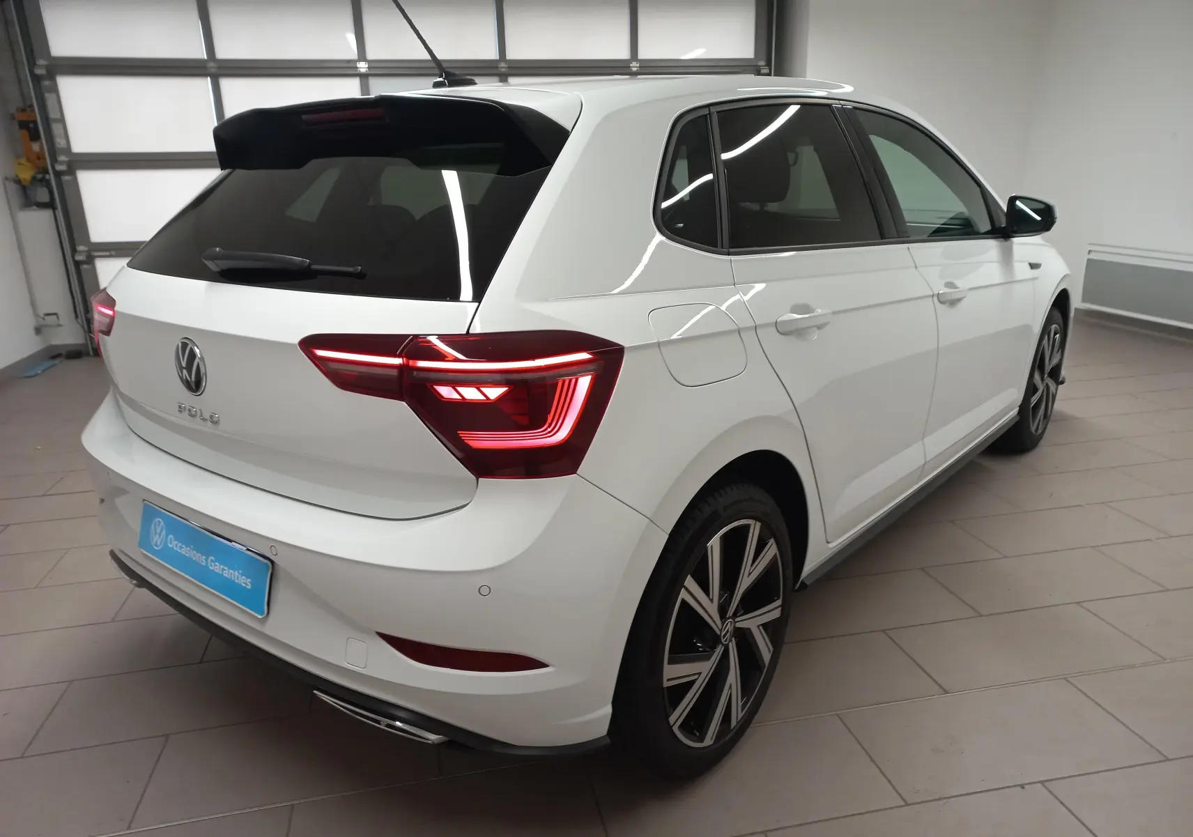 Vue 3/4 arrière droite d'une Volkswagen Polo blanche avec vitres arrière surteintées et feux arrière allumés en intérieur.