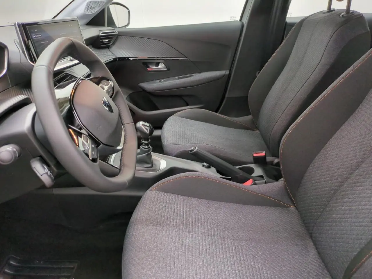 Vue intérieure côté conducteur de la Peugeot 208 2025, sièges tissu gris avec surpiqûres orange et volant cuir compact.