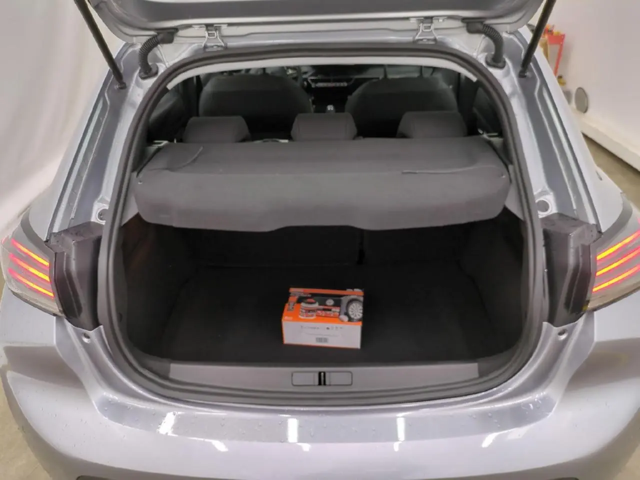 Coffre ouvert vu de l'arrière d'une Peugeot 208 gris clair, avec cache-bagages et boîte orange à l'intérieur.