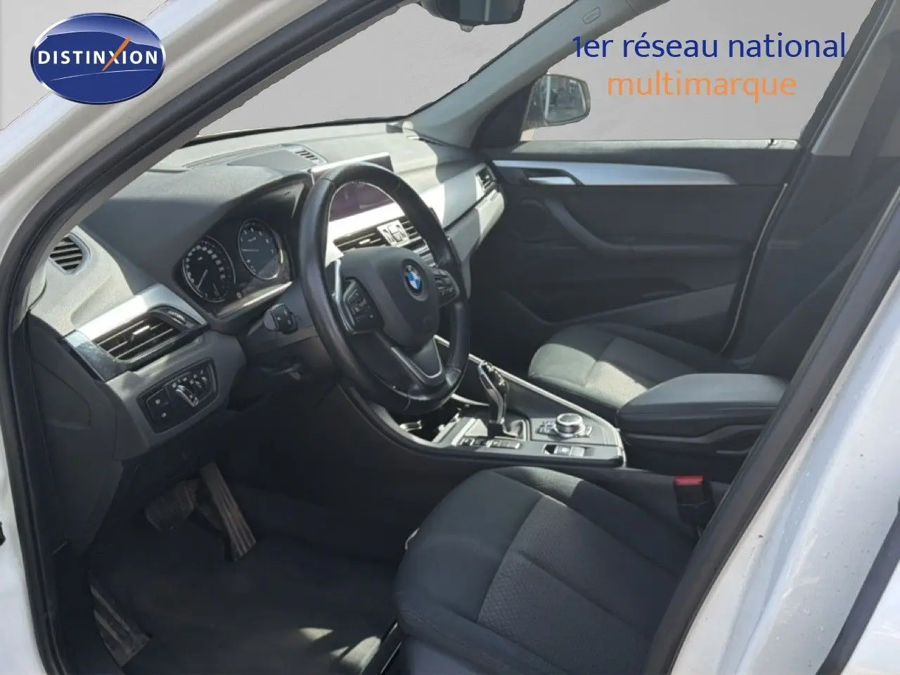 Vue intérieure côté conducteur d'un BMW X1 blanc 2022, volant multifonction et sièges tissu noirs visibles.