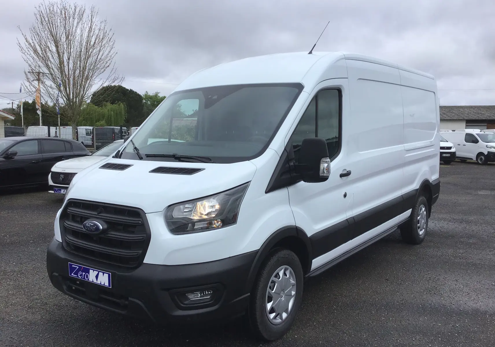 Fourgon blanc Ford Transit 350 L3H2 vu en 3/4 avant droit avec calandre noire et phares allumés.