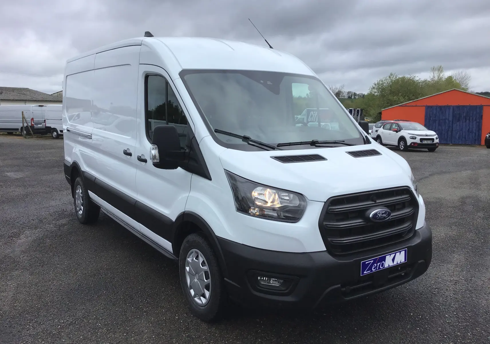 Ford Transit Fourgon blanc vu en 3/4 avant droit avec calandre noire et phares allumés sur parking extérieur.