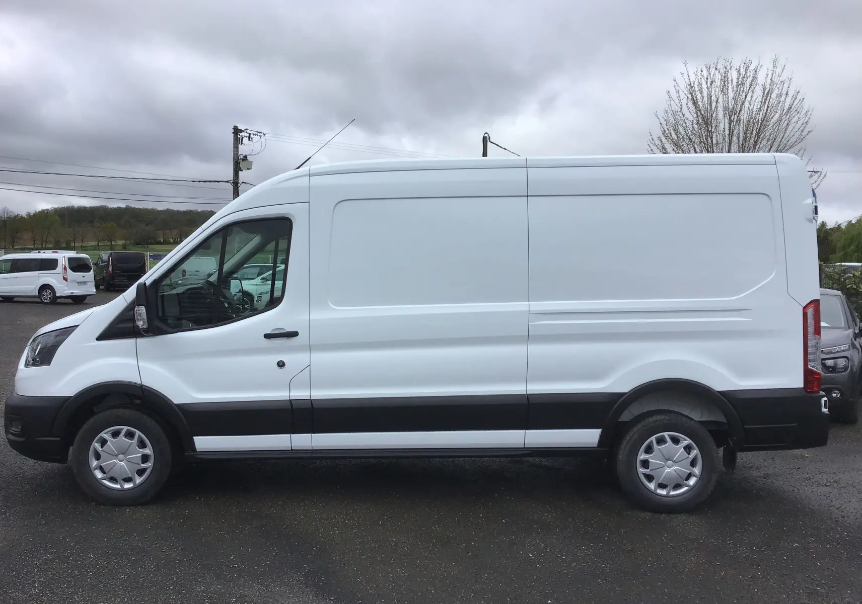 Profil côté gauche d'un fourgon Ford Transit blanc 350 L3H2 2023 avec jantes acier et bandeau noir sur bas de caisse.