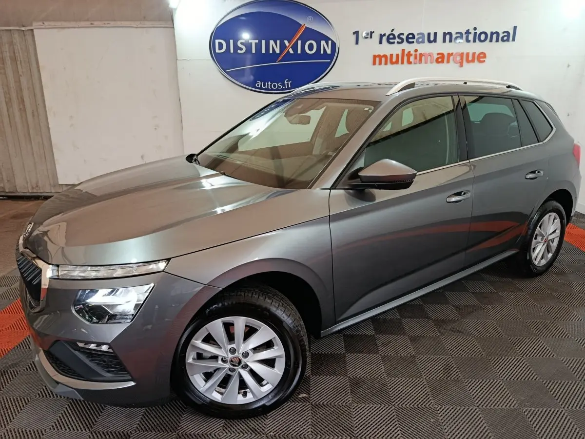 Vue 3/4 avant droit d’un Skoda Kamiq gris clair avec jantes alliage et barres de toit blanches en intérieur showroom.